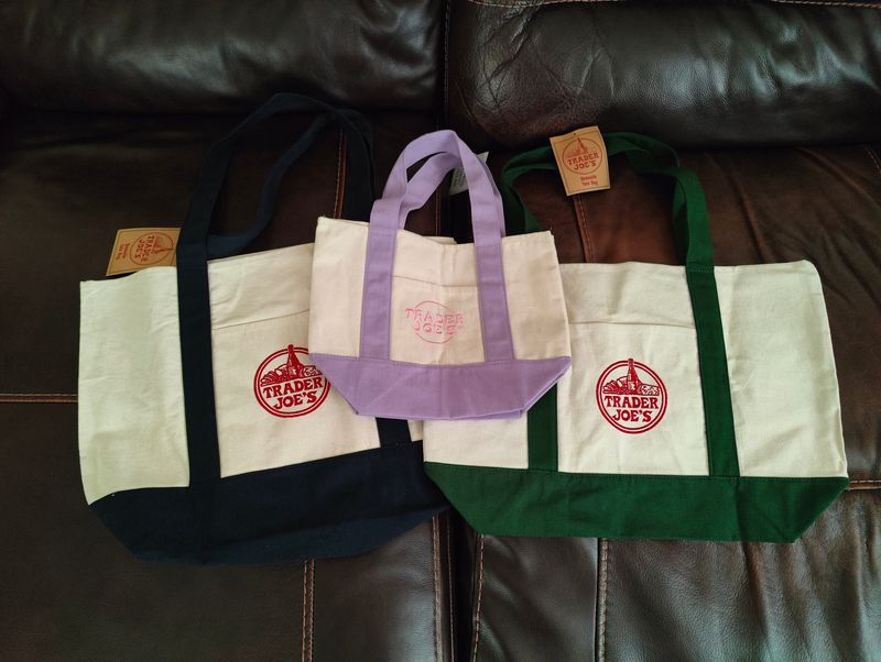 A Trader Joe's lavender pastel mini canvas tote bag lays atop two large canvas tote bags with blue and green trim. A large version of the lavender tote was the latest design announced by Trader Joe's.