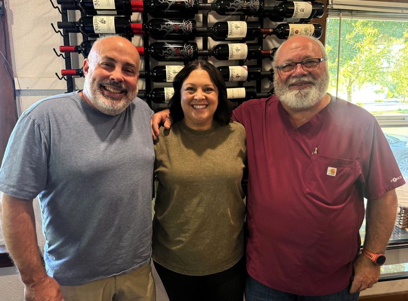 The Grape Escape owners Adam Browning and Kayla Hatch pose for a photo inside their downtown Redding wine bar on Aug. 1, 2026, alongside Jim Farmer, right, who offers a food menu for customers.
