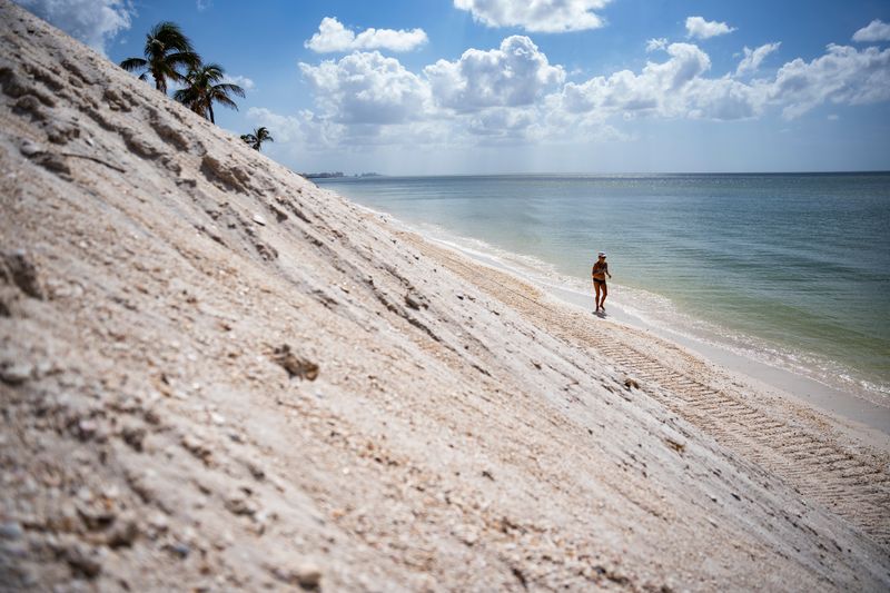 A beachgoer walks near piles of sand near Barefoot Beach in Naples as Collier County continued a beach regrading project on Thursday, Oct. 31, 2024.