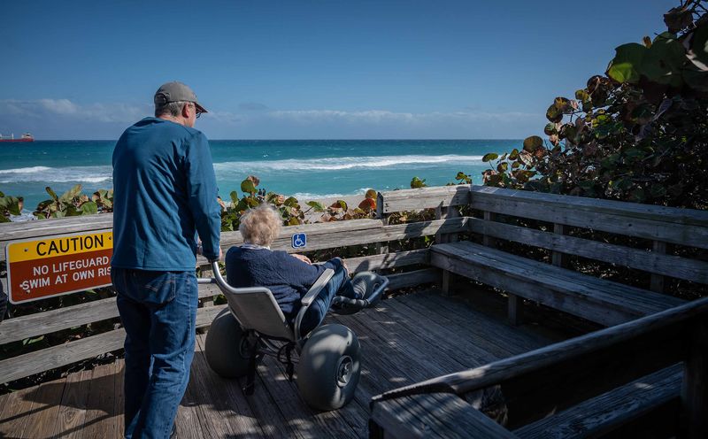 David Muse, Carmel, Ind., pushes his mother-in-law Beverly Schmidt, Columbus, Ohio, southeast on the boardwalk to a parked viewing position of the Atlantic Ocean shoreline on a beach wheelchair, available at no charge on a first-come, first-served basis, at John D. MacArthur Beach State Park in North Palm Beach, Fla., on January 8, 2025.