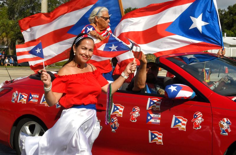 United Third Bridge's "Godmother of the Year" Ada Garcia has a seat of honor in a convertible during the 28th annual Puerto Rican Day parade in Palm Bay, while Maria Camacho, a joyful parade participant, waves a flag alongside the car.