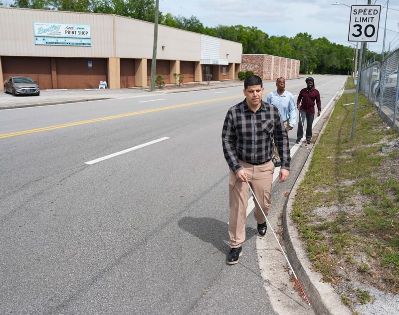 Center for Independent Living Jacksonville employees Jose Morales, foreground, Jermaine Raymer, center and Travis Brown, right, all of whom are legally blind, make their way to the Sandwich House for lunch along Art Museum Drive without the safety of a sidewalk Monday afternoon, April 13, 2026. . [Doug Engle/Florida Times-Union]