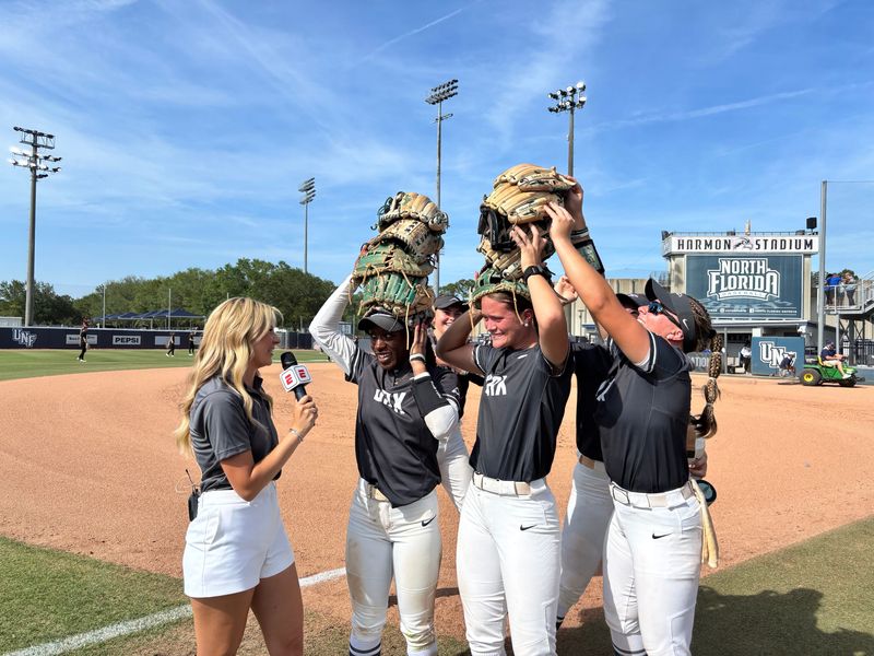 Jacksonville University softball team members playfully pile gloves on the heads of teammates Jaida Thomas (center) and Jacey Harrelson (right) while they were interviewed on ESPN+ by Cecily Webb after the Dolphins beat Florida Gulf Coast 6-2 on May 6 in the ASUN Softball Tournament, at the University of North Florida Softball Complex.