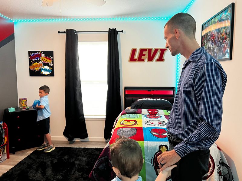 Levi Bowles, 6, left, sees his room for the first time June 3, 2026, at the new home his father, Garrett Bowles, right, financed with the help of Habitat for Humanity, a national affordable housing nonprofit. The superhero-themed décor was sponsored by the iTHINK Community Foundation that furnishes and decorates the house of one affordable housing applicant per year as part of their “build up” initiative.