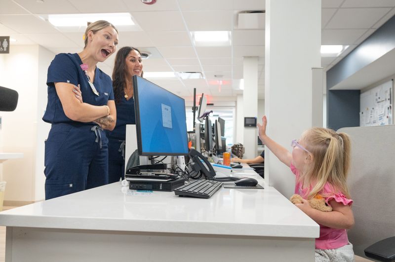 From right, Baylee Vaughn, 4, waves to register nurses Sara Carroll and Mallarie Rigby while sitting at their desk at the new Ascension Sacred Heart Emergency Department in Pace on July 28, 2026. The ED medical team helped save the then 3-year-old Baylee’s life when she almost drowned in a swimming pool on June 13th.