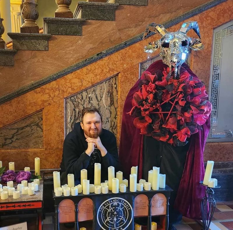 Mortimer Adramelech, a minister with The Satanic Temple of Iowa, poses with a display the group erected in the Iowa Capitol in 2023. The display was later vandalized.