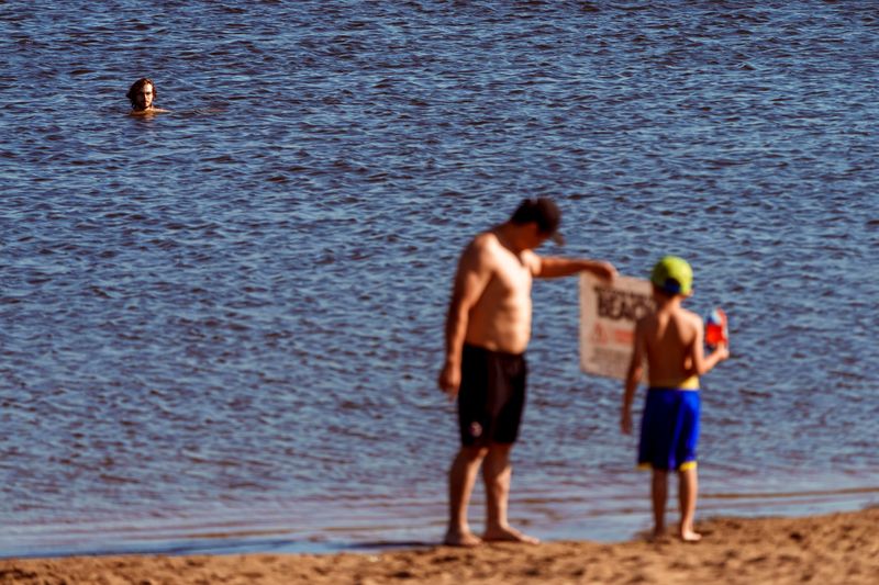 People read a loose warning sign that was blown across the beach at the Blue Heron Lake in Raccoon River park Wednesday, June 17, 2026 in West Des Moines. Some people continue to swim despite the warning signs not recommending swimming due to high E.coli in the water.