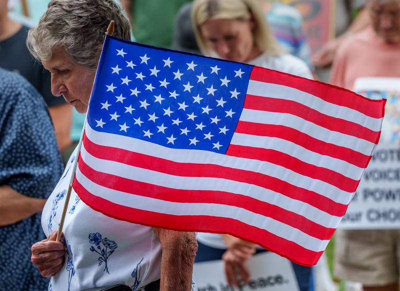 An American flag held by a protester flies in the breeze during a pre-march prayer Thursday, July 17, 2025 at John Gwynn Park in Peoria. The march was part of the nationwide â€œGood Trouble Lives Onâ€ protests inspired by late civil rights activist John Lewis.
