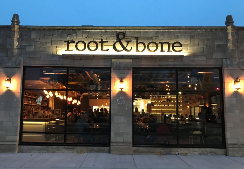 At twilight, under a deep-azure sky, lanterns and backlights illuminate buff stonework framing the warm, golden bustle of diners inside Root & Bone. The restaurant opened in January 2020 at 4601 N. College Ave., south of Broad Ripple in Indianapolis.