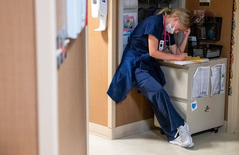 Tired, a nurse sits propped against a wall Monday, Sept. 20, 2021, inside Franciscan Health's critical care unit. She's filling out paperwork as fellow nurses and doctors hold their rounds.