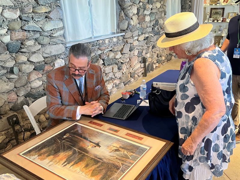 Nicholas Lowry, who often appraises posters on Antiques Roadshow while wearing spectacular suits, takes a look at the print "Autumn Float" by Jim Foote brought by Karla to the show in Charlevoix on July 1, 2025. "I've watched you for years," Karla told Lowry.