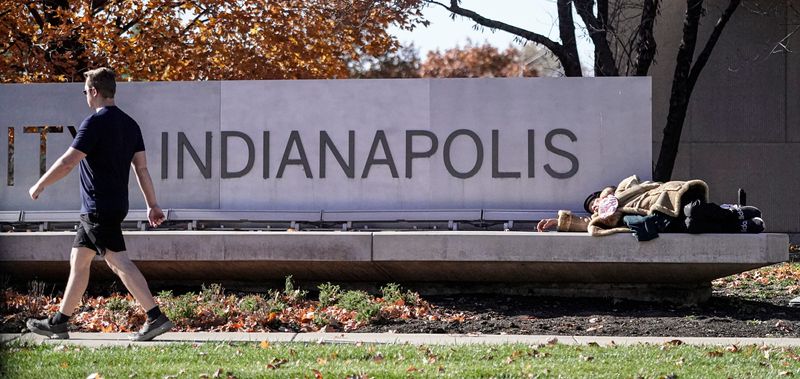 An elderly woman wearing two jackets on a brisk day, sleeps on an Indiana University sign along West Street on Thursday, Nov. 13, 2025, in Indianapolis.