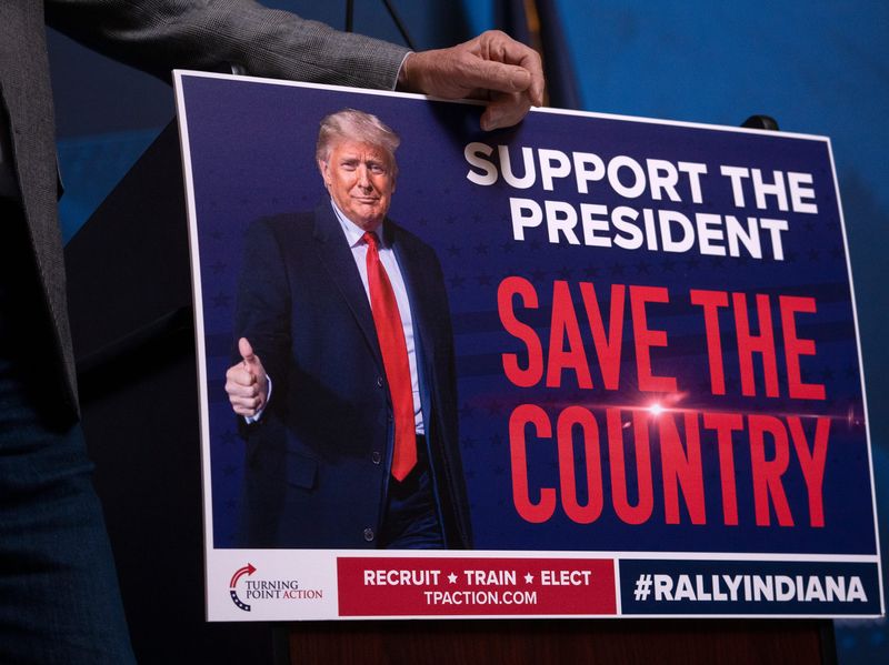 Gov. Mike Braun leans on a pro-Trump sign as he addresses the crowd during the All In For Indiana gathering, a pro-redistricting rally, Friday, Dec. 5, 2025 at the Indiana Statehouse.