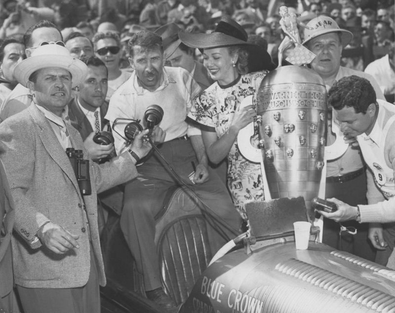 The Borg-Warner trophy is presented to three time Indy 500 winning driver (second win in 47) Mauri Rose by actress and USO star, Carole Landis in victory lane on May 30, 1947, at the Indianapolis Motor Speedway. Wilbur Shaw, Speedway president is in the white hat. Carole Landis was dubbed the “Ping Girl” after a popular motor oil ad at the time that claimed to take the ‘ping’ out of an engine and “make it purr.” She died under suspicious circumstances in 1948.