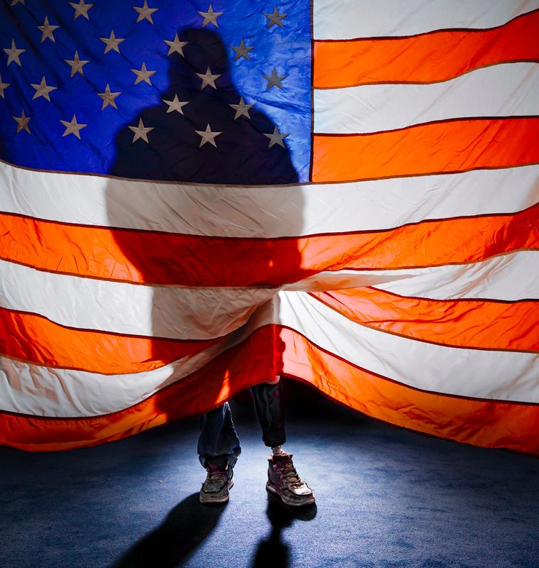 A man who sought asylum in the U.S. in 2024 lifts the bottom half of an American flag to reveal his amputated left leg. The father of four and manual laborer told IndyStar that his left leg was amputated after wearing an ankle monitor outfitted by BI Incorporated, a private government contractor that provides monitoring services to ICE. He said the device was too tight and caused swelling and infection.