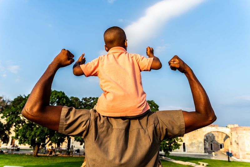 Rear view of a father with his son flexing muscles outdoors.