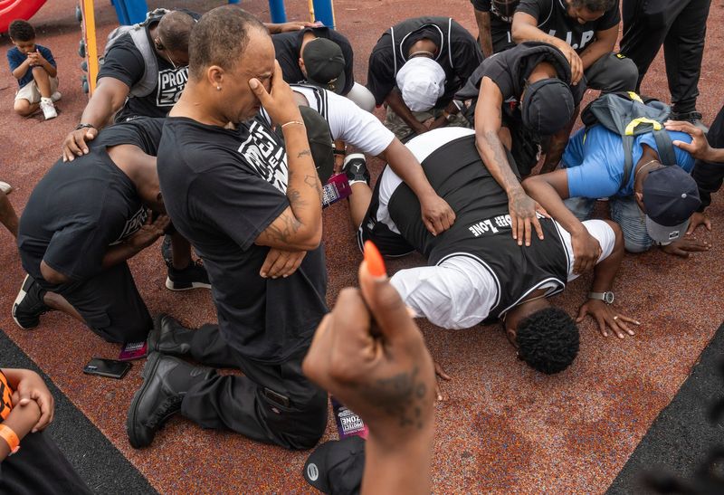 Team Pursuit member Phil Sample, left, of Detroit, takes a knee, as Pastor Mo Hardwick of Live in Peace Movement, right, leads others from Detroit’s Community Violence Intervention Organizations to get on the ground at Skinner Playfield next to Denby High School on Detroit’s east side on Monday, June 30, 2025, in prayer at the scene where a triple shooting at the park occurred on Friday that killed 4-year-old Samir Josiah Grubbs.