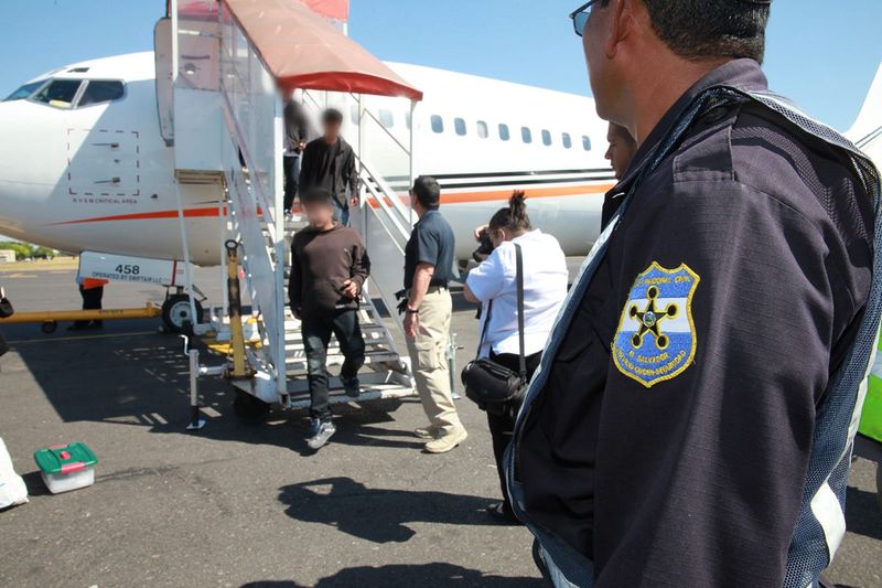 People deported by ICE step off a flight in El Salvador, as a Salvadoran national guard member looks on.