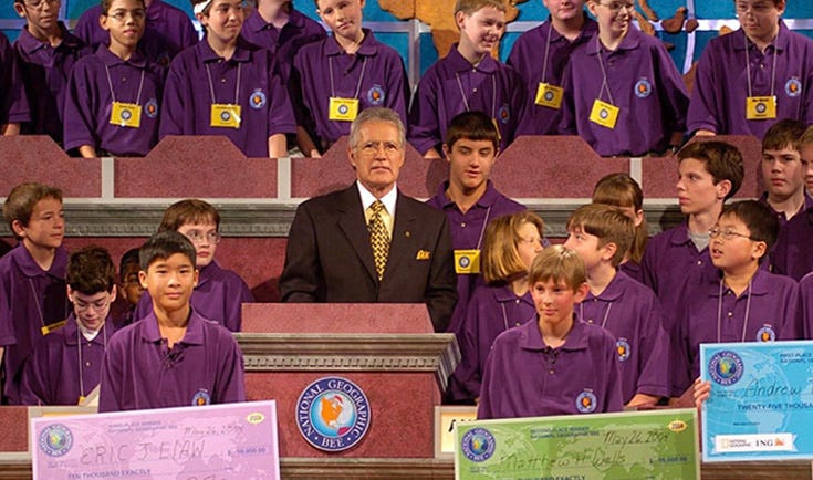 Alex Trebek stands behind a podium at the 2004 National Geographic Bee in Washington DC, as then-middle school student Jamie Ding, standing on far right, looks on.