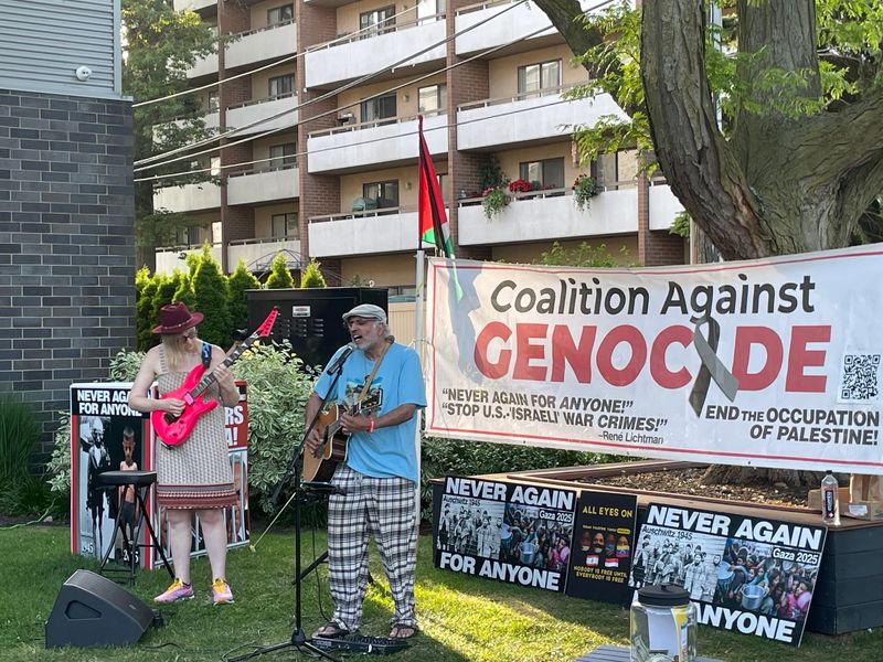 Amrit Kohli, founder of Queer Folk, sings at a concert at Schiffer Park in Ferndale, Michigan, on June 27, 2026, that was a benefit for the Michigan Coalition Against Genocide, which advocates for Palestinians. Accompaying him on guitar is Ally Fies, to his right. A Palestinian flag waves behind him along with anti-war signs and posters reading "Never Again."