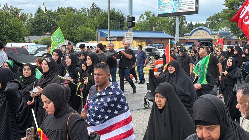 Arbaeen procession is seen here on Greenfield Road in Dearborn, Michigan, on Aug. 2, 2026. A man has an American flag wrapped around him. Many are dressed in black as a symbol of mourning as they recall the death of Imam Hussain in the 7th century.