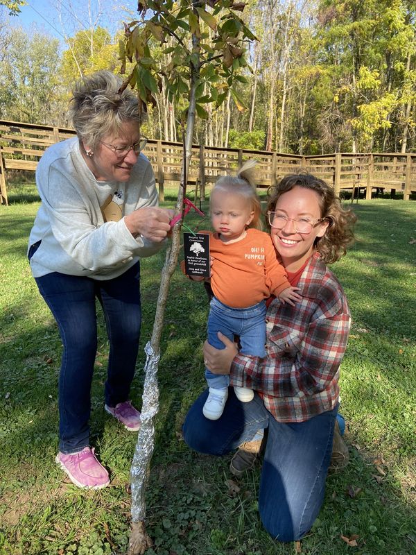 June Gebhardt attaches a tag to an Ohio buckeye tree honoring the birth of her first grandchild, Sophie, who looks on with mother Sara Rowlinson near the Friendship Bridge in Aumiller Park.