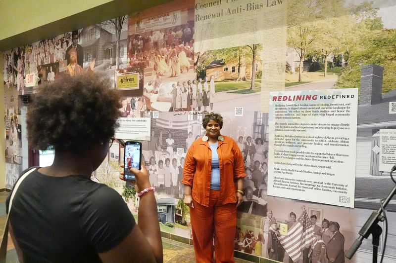 Toni Harper takes a photo of her mother, Dara Harper, founder of Akron Black Artist Guild and the artist who created the mural “Redlining Redefined,” before the official unveiling of the work in the Akron Urban League on June 20, 2026.