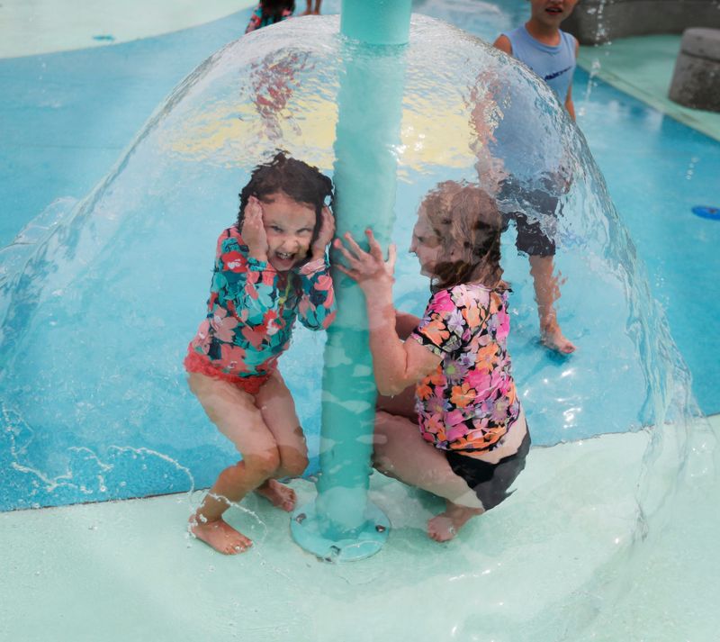 Three-year old Adaleigh Johnson and her 9-year-old sister Braleigh Johnson stand under cascading water Monday, July 8, 2024 at Rodgers Park. The City of Lubbock Parks and Recreation held three “Splash Around Town” events Monday to show off their three new splash pads.