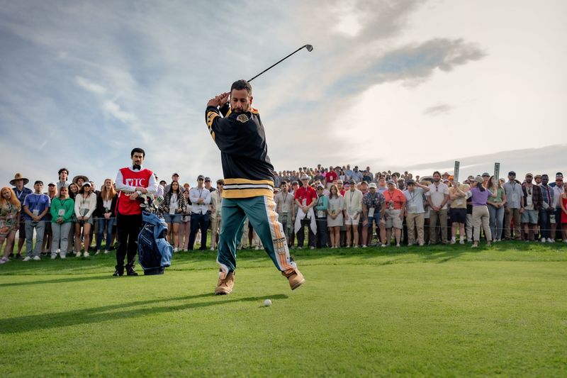 Happy Gilmore, played by Adam Sandler, breaks out his signature golf swing as caddie Oscar, played by Bad Bunny, looks on in "Happy Gilmore 2," released to stream on Netflix early July 25, 2025.