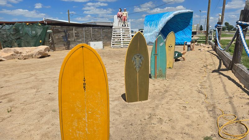 Colorful surfboards line a sandy photo area at Shark Beach, with a wave backdrop and mannequin lifeguards creating a playful vacation vibe in Amarillo.