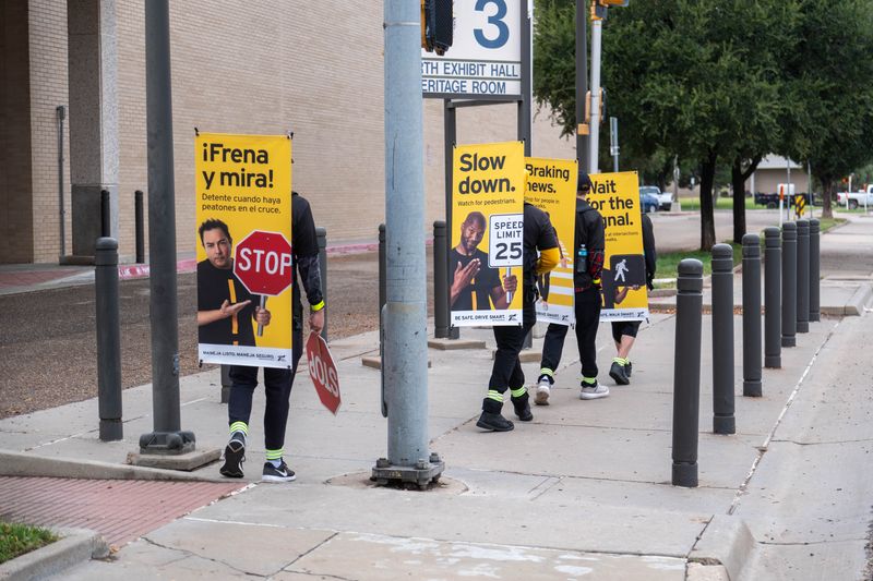 Participants in TxDOT’s “Be Safe. Drive Smart.” campaign line up at a downtown intersection, displaying messages like “Slow down,” “Wait for the signal,” and “¡Frena y mira!” during Pedestrian Safety Month.
