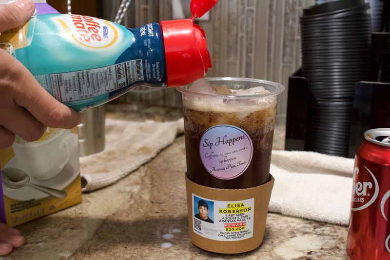 A barista at Sip Happens in Aransas Pass starts to pour creamer in a dirty soda with a cup sleeve featuring Elisa Roberson on Aug. 4, 2026. Elisa went missing in 1989 and was legally declared deceased in 2025.