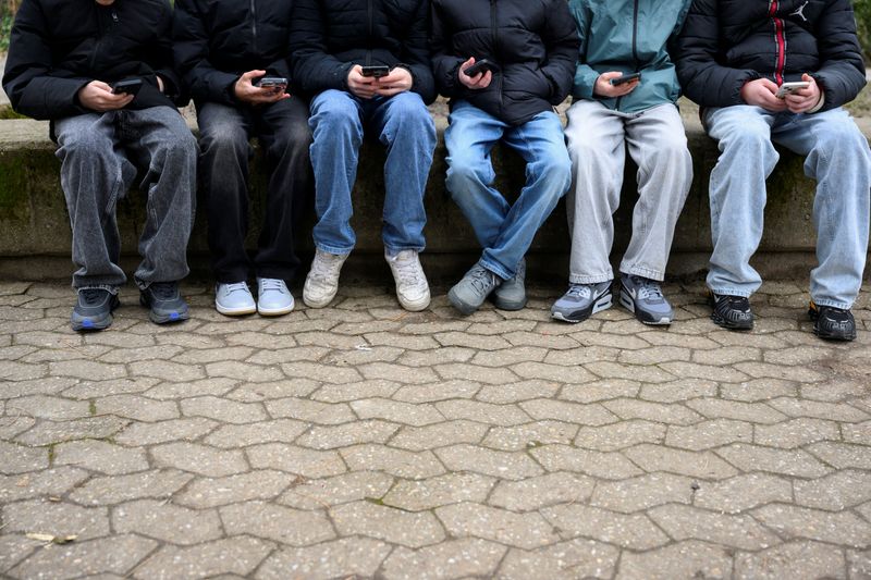 Teenagers pose for a picture while looking at their phones. February 20, 2026. REUTERS/Jana Rodenbusch