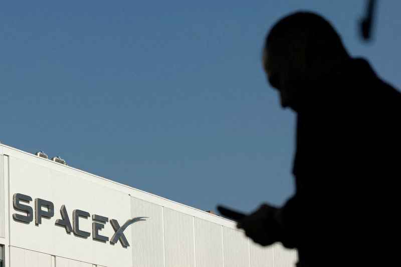 FILE PHOTO: SpaceX logo as an employe looks at his phone while making his way to work at the company’s facility on the day of the SpaceX IPO, in Hawthorne, California, U.S. June 12, 2026.  REUTERS/Mike Blake/File Photo