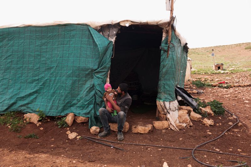 Suhaib Abu Kbash sits with his child on his lap outside their makeshift house in a Palestinian Bedouin community in the Jordan Valley, in the Israeli‑occupied West Bank, March 19, 2026. REUTERS/Mohammed Torokman