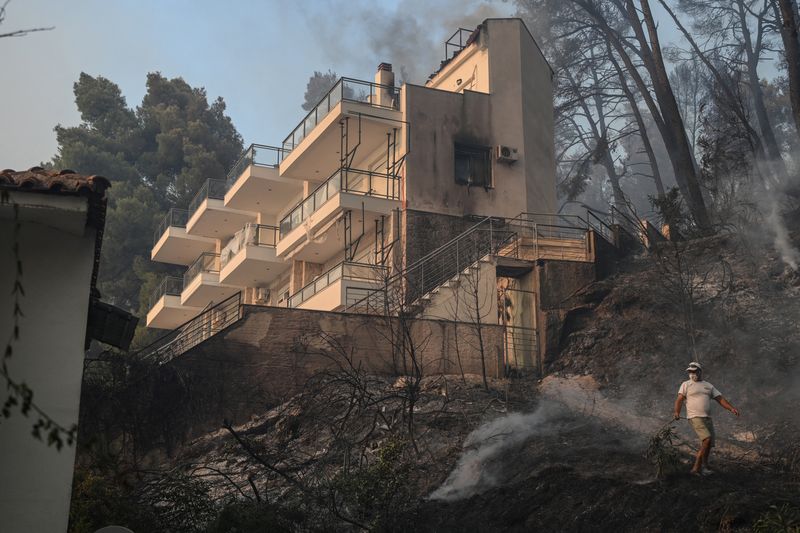A man walks past a house during a wildfire fanned by strong winds at the resort of Siviri on the Halkidiki peninsula, Greece, August 13, 2026. REUTERS/Alexandros Litsardakis