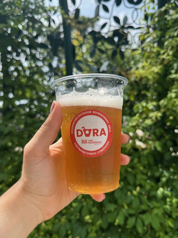 A beverage in a clear cup sporting an official DORA sticker. Patrons are now able to take their drinks outside within the bounds of downtown Manitowoc.