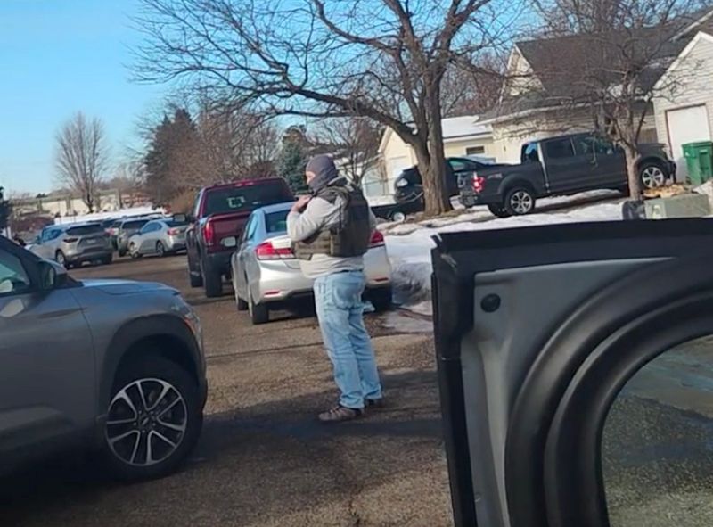 A masked immigration enforcement agent stands in the road during an apparent operation in Hudson, Wisconsin. This still image comes from a video taken by Hudson resident Addison Filiatreaux, who helps coordinate donations for immigrant families. The video shows several vehicles leaving a neighborhood while the pictured agent blocks the road.