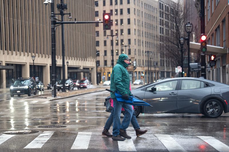 Two pedestrians cross the street holding their retracted umbrella, while high winds cause others to struggle with theirs in the rain, on the afternoon of March 15, 2026 in Milwaukee, Wisconsin.