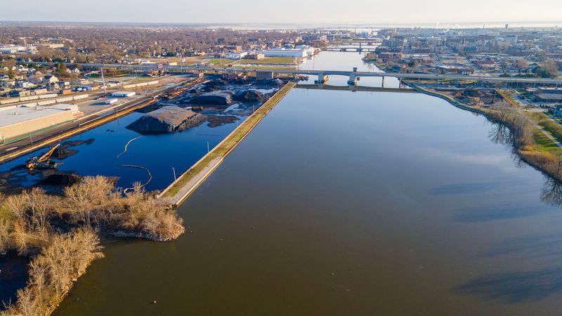 Piles of coal (left) are seen the lower Fox River south of the East Mason Street Bridge in Green Bay, Wisconsin on April 23, 2026. A $1.3 billion PCB cleanup remediation effort in the river spanned about 17 years and concluded in 2020 after removing roughly 6.5 million cubic yards of contaminated sediment, with the EPA certifying the cleanup as complete in 2023 and long-term monitoring continuing for decades.