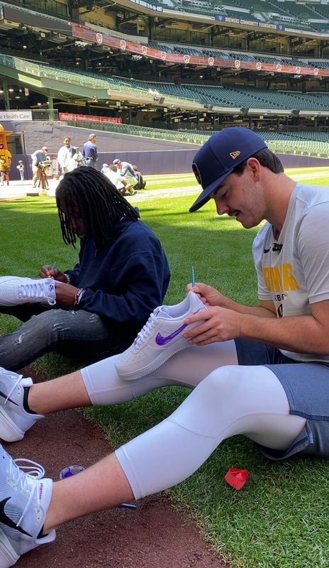 Milwaukee Brewers starting pitcher Logan Henderson and 14-year-old Shane Cox sitting on the edge of the grass along the warning track of American Family Field, painting the swooshes on their new white Nike Air Force 1s purple on Tuesday, June 2, 2026.